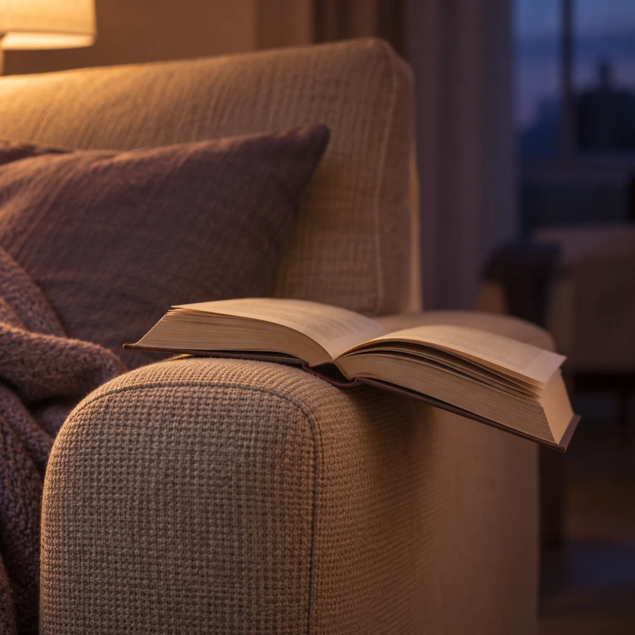 Un livre ouvert posé sur l’accoudoir d’un canapé, à la lumière chaude d’une lampe, le soir